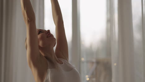A-young-woman-in-white-sportswear-is-stretching-with-a-large-hall-with-large-windows-in-a-slow-motion-scheme-the-sun's-rays-shine-through-the-window.